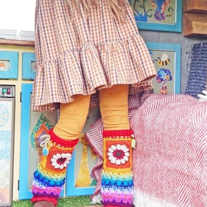 May include: A person wearing a brown and white checked dress, mustard-coloured leggings, and brown shoes. The person is wearing colourful, rainbow-striped crochet leg warmers with a white flower design. The background includes framed art and a red and white blanket.