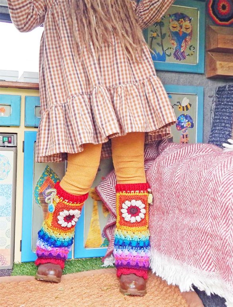 May include: A person wearing a brown and white checked dress, mustard-coloured leggings, and brown shoes. The person is wearing colourful, rainbow-striped crochet leg warmers with a white flower design. The background includes framed art and a red and white blanket.