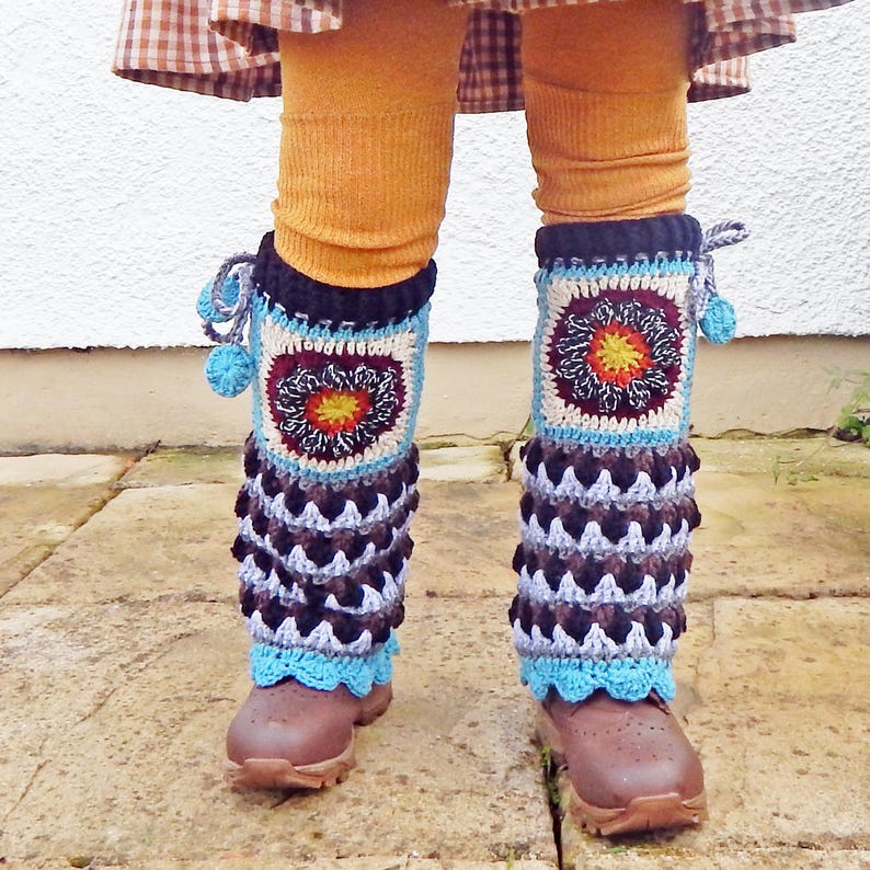 May include: Handmade crochet leg warmers in brown, blue, and cream. The leg warmers have a square design with a central floral pattern and a scalloped edge. Small blue pom-poms are attached to the top.