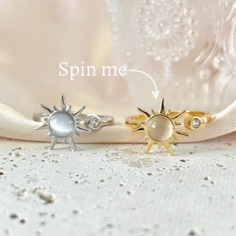 May include: Two rings with sunburst designs. One ring is silver-toned, the other is gold-toned. Both rings have a central, round, light-colored stone and a smaller, clear stone. The text "Spin me" is above the rings.