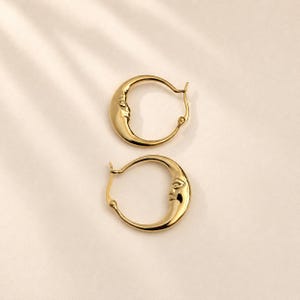 Op de afbeelding: Een paar gouden oorringen met een halve maan ontwerp, voorzien van een gedetailleerd gezicht in reliëf aan de zijkant van elke ring. De oorbellen hebben een gepolijste metalen afwerking en een scharniersluiting voor bevestiging aan de oorlel.