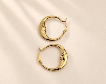May include: A pair of gold hoop earrings featuring a crescent moon design with a detailed face embossed on the side of each hoop. The earrings have a polished metallic finish and a hinged closure mechanism for secure attachment to the earlobe.