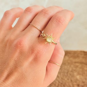 May include: A hand wearing two gold rings. One ring features a delicate sunburst design with a central, pale gemstone. The other ring is a simple, thin band. The rings are set against a neutral background.