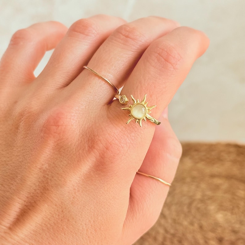 May include: A hand wearing two gold rings. One ring features a delicate sunburst design with a central, pale gemstone. The other ring is a simple, thin band. The rings are set against a neutral background.