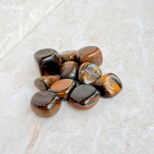 May include: A collection of polished tiger's eye stones. The stones vary in size and shape, with a mix of dark brown, golden brown, and black colors. The surface of each stone is smooth and reflective, with visible chatoyancy.