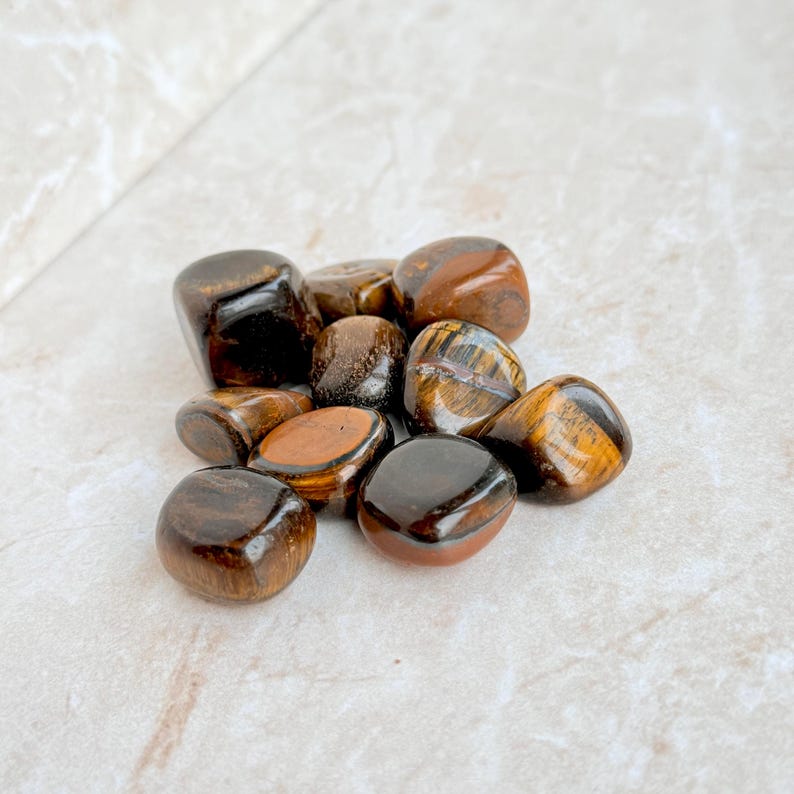 May include: A collection of polished tiger's eye stones. The stones vary in size and shape, with a mix of dark brown, golden brown, and black colors. The surface of each stone is smooth and reflective, with visible chatoyancy.