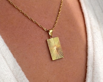 May include: A gold-colored necklace with a rectangular pendant. The pendant has a sunburst design with vertical lines radiating from the center. The chain is a twisted rope style, and the necklace is worn against a white fabric.