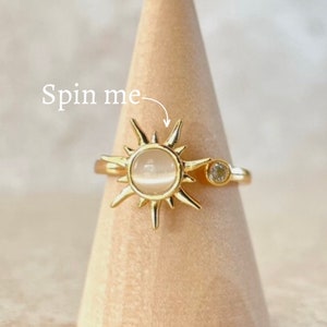 May include: Gold ring with a spinning sun design featuring a white stone and a small clear stone accent. The text "Spin me" is written on the image.