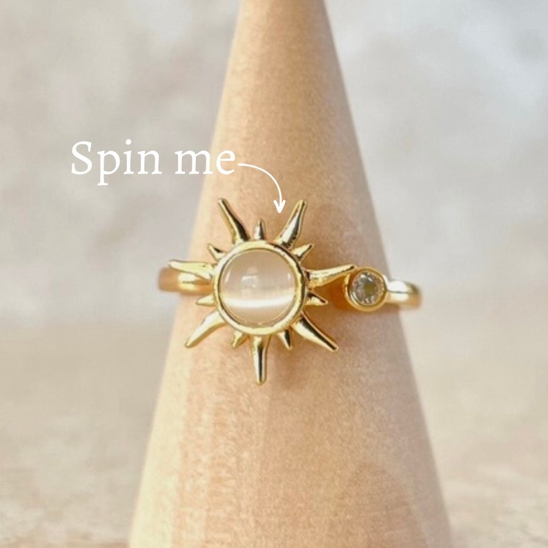 May include: Gold-toned ring featuring a sunburst design with a central, round, light-colored stone and a smaller, clear stone accent. The ring is displayed on a wooden cone with the text "Spin me" in white.