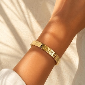 May include: A gold-toned cuff bracelet featuring a textured, hammered surface with an irregular, organic pattern. The metal band has a polished finish that reflects light, creating a bright, metallic appearance across the entire outer surface.