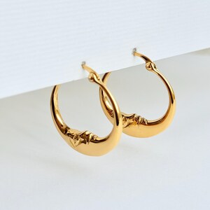 May include: A pair of gold-toned hoop earrings featuring a crescent moon design with a sculpted face. The earrings have a polished metallic finish and a hinged closure mechanism, showcasing a smooth, curved silhouette that forms a circular shape.