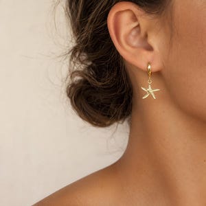 May include: A gold hoop earring featuring a small, textured starfish charm dangling from the bottom. The earring is crafted from a polished gold-tone metal, showcasing a detailed five-pointed starfish design that hangs freely from the circular hoop.