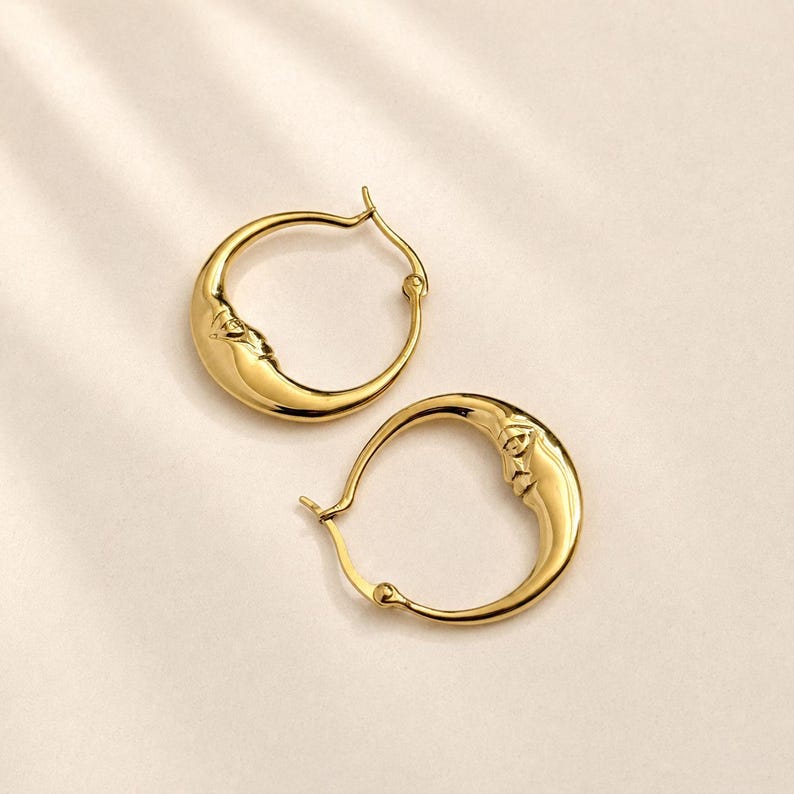 May include: A pair of gold hoop earrings featuring a crescent moon design. Each hoop is shaped like a curved moon with a detailed, embossed face on the side. The earrings have a polished, reflective finish and a simple hinged closure mechanism.