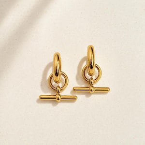 May include: A pair of gold-toned earrings featuring a polished hoop connected to a circular ring, which suspends a horizontal bar toggle component. The earrings have a smooth, reflective metallic finish and a structured, geometric design.