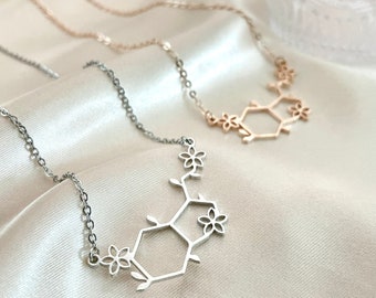 May include: Two necklaces with floral and geometric pendant designs. One necklace is silver-toned, and the other is rose gold. The pendants feature a hexagonal shape with floral accents, suspended from delicate chains. The necklaces rest on a satin surface.