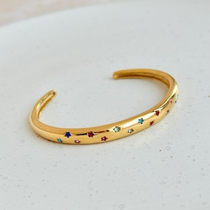 Op de afbeelding: Goudkleurige manchetarmband met kleurrijke stervormige accenten. De open armband heeft rode, blauwe, groene en roze sterren. De sieraden zijn geplaatst tegen een witte achtergrond.