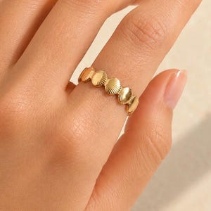 May include: A gold-toned ring featuring a continuous band of small, repeating shell-shaped motifs. The metal has a polished, reflective surface that catches the light, creating a textured appearance across the entire circumference of the ring.