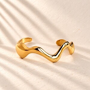 May include: A gold-toned cuff bracelet featuring a continuous, fluid, and wavy design with a polished, reflective surface. The open-ended band has rounded, smooth terminals and a thick, organic shape that curves along its entire length.