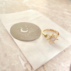 May include: Gold-colored adjustable ring featuring a sunburst design with a central white stone and a small clear stone on the opposite end. A small round sticker with a crescent moon, stars, and hands is also visible.