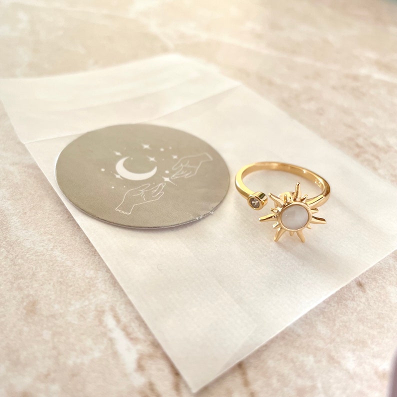 May include: Gold-colored adjustable ring featuring a sunburst design with a central white stone and a small clear stone on the opposite end. A small round sticker with a crescent moon, stars, and hands is also visible.