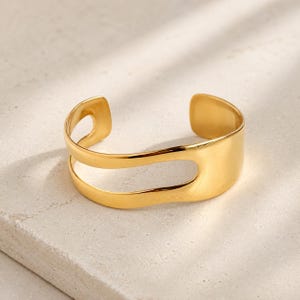 May include: A gold-coloured cuff bracelet with a distinctive cutout design. The open-ended bracelet has a smooth, polished finish and a modern, minimalist aesthetic. The bracelet is a stylish accessory.