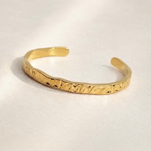 May include: A gold-toned cuff bracelet featuring an open design and a textured, hammered surface. The metal band has an irregular, organic shape with a polished finish that reflects light across its uneven, indented exterior surface.