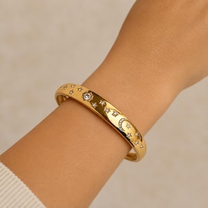 May include: A gold-toned bangle bracelet featuring a polished finish, adorned with a series of small, embedded clear stones arranged in celestial patterns, including multiple star shapes and a crescent moon motif along the outer surface.