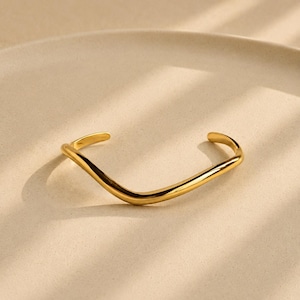 May include: A polished gold-tone cuff bracelet featuring a smooth, continuous wave-like design. The metal surface is reflective and curved, forming an open-ended circular shape that tapers slightly at both ends, creating a fluid and organic silhouette.