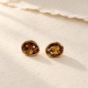 May include: A pair of stud earrings featuring oval-shaped, faceted brown gemstones set in polished gold-tone metal frames. The stones exhibit a deep, translucent amber hue with intricate geometric cuts that reflect light across the surface.