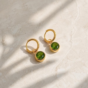 May include: A pair of gold hoop earrings, each featuring a single round, faceted, vibrant green gemstone charm suspended from the bottom of the hoop. The earrings have a polished metallic finish and a classic circular design.