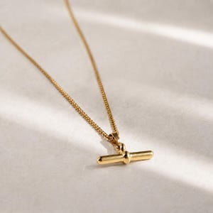 Op de afbeelding: Een goudkleurige ketting met een T-bar hanger. De fijne ketting leidt naar een staafvormige hanger met een knoopdetail. De ketting wordt gepresenteerd op een wit oppervlak, met zachte verlichting.
