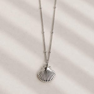 May include: A silver-toned necklace featuring a delicate chain with small, evenly spaced metal beads and a pendant shaped like a classic scalloped seashell. The pendant has a polished, reflective surface with vertical ridges radiating from the top.