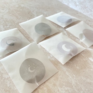 May include: Six square translucent white paper envelopes, each containing a circular sticker. The stickers feature various celestial designs, including crescent moons and stars in muted grey and beige tones, visible through the semi-transparent paper.