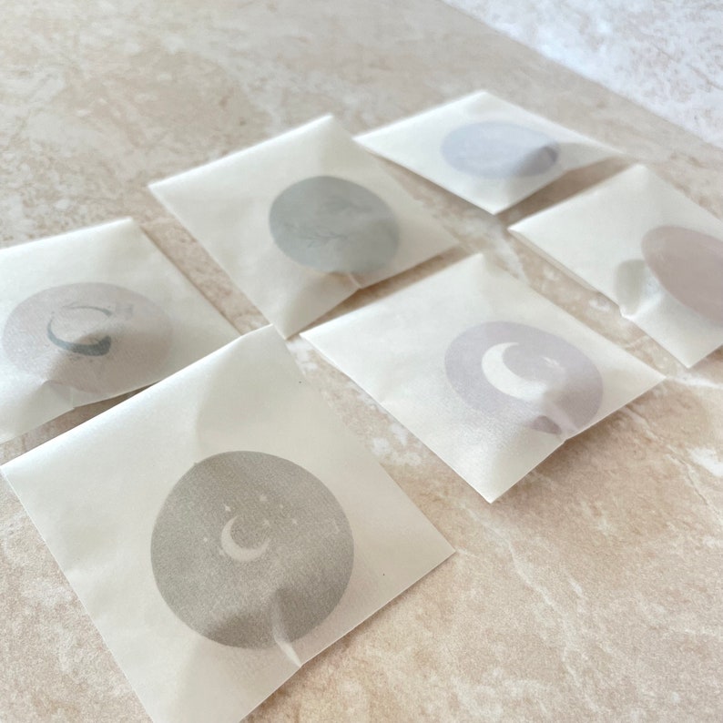 May include: Six square translucent white paper envelopes, each containing a circular sticker. The stickers feature various celestial designs, including crescent moons and stars in muted grey and beige tones, visible through the semi-transparent paper.