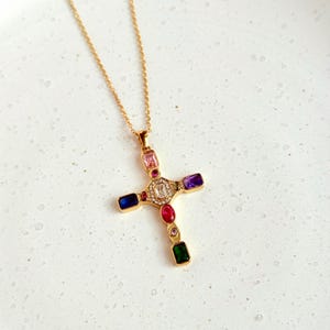 May include: Gold necklace featuring a cross pendant embellished with colourful gemstones. The cross incorporates pink, purple, red, green, and blue stones, with a central clear stone surrounded by smaller clear stones.