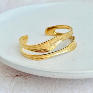 May include: A gold-colored cuff bracelet with a textured, leaf-like design. The open-ended bracelet features two overlapping bands that curve around the wrist. The jewelry is displayed on a white plate.