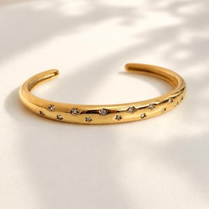 May include: A gold-toned open cuff bracelet featuring a polished finish and a series of small, star-shaped cutouts along the outer band. Each star cutout is set with a single, clear, sparkling gemstone, creating a celestial-inspired design.