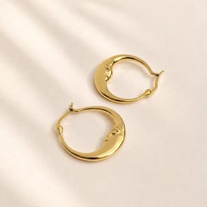 May include: A pair of gold hoop earrings, each featuring a sculpted crescent moon face design. The earrings have a polished, reflective finish and a hinged closure mechanism, showcasing a circular shape with a detailed celestial profile.
