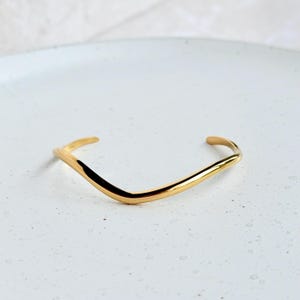 Op de afbeelding: Een goudkleurige, V-vormige manchetarmband. Het minimalistische ontwerp heeft een gladde, gebogen band die taps toeloopt aan de uiteinden. De armband wordt gepresenteerd op een wit oppervlak, wat de gepolijste afwerking benadrukt. Een eenvoudig, elegant sieraad.