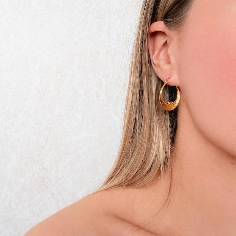 May include: A gold hoop earring with a smooth, polished finish. The earring features a circular design that tapers slightly at the bottom, creating a subtle crescent shape. The metal has a warm, reflective yellow tone and a simple, clean structure.