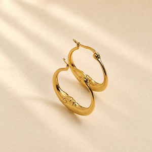 May include: A pair of gold-toned hoop earrings featuring a smooth, polished finish and a curved, crescent-like shape. The earrings have a hinged clasp mechanism at the top, allowing them to open and close securely for wear on the earlobes.
