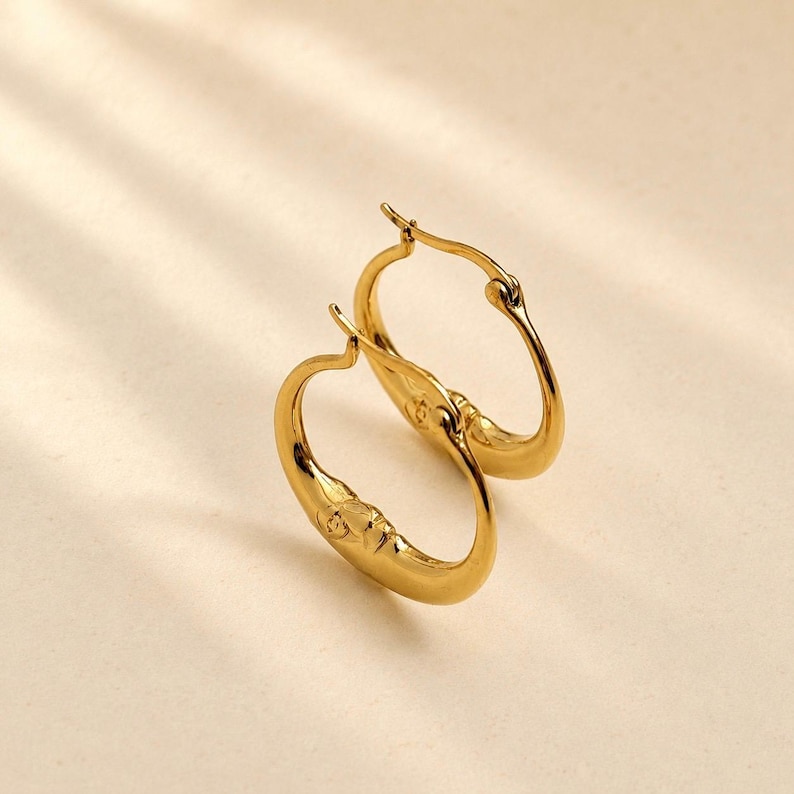 May include: A pair of gold-toned hoop earrings featuring a smooth, polished finish and a curved, crescent-like shape. The earrings have a hinged clasp mechanism at the top, allowing them to open and close securely for wear on the earlobes.