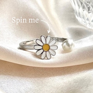 May include: A silver ring featuring a white daisy flower and a pearl detail. The ring is designed to spin. The text "Spin me" is written on the image.