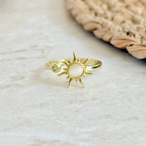 May include: Gold-colored ring featuring a sunburst design with a central, round, off-white stone. The ring has a delicate band and a smaller, clear stone on the side. The ring is set against a neutral background.
