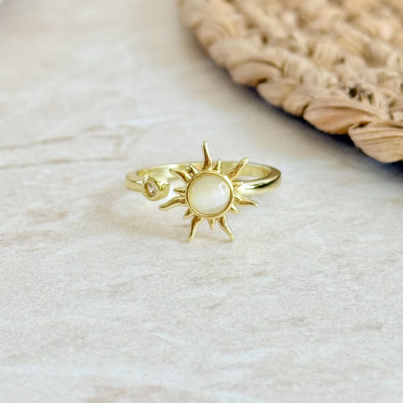 May include: Gold-colored ring featuring a sunburst design with a central, round, off-white stone. The ring has a delicate band and a smaller, clear stone on the side. The ring is set against a neutral background.