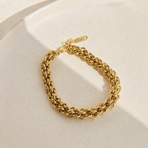 May include: A gold-toned chain bracelet featuring a thick, interlocking woven link design. The bracelet includes a small, rectangular metal tag attached near the clasp, which displays engraved text that is partially visible.