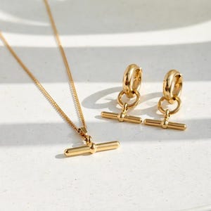 May include: A gold-toned jewellery set consisting of a necklace and a pair of hoop earrings. The necklace features a delicate chain with a horizontal bar pendant. Each hoop earring includes a matching small horizontal bar charm suspended from the ring.