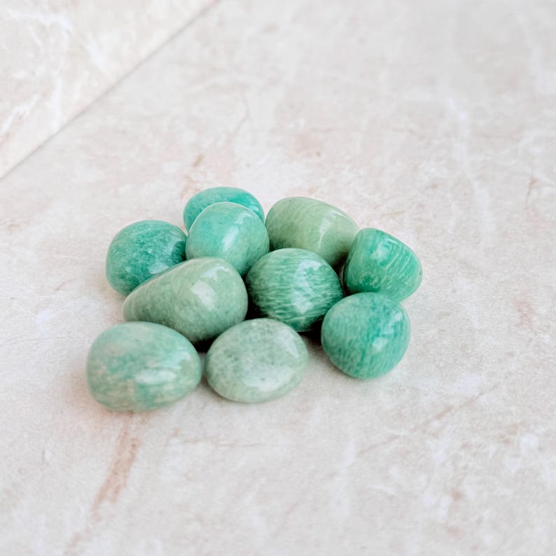 May include: A collection of smooth, polished Amazonite stones in varying shades of green and teal. The stones are oval and rounded, with a natural, earthy appearance. They are arranged in a loose pile, suggesting a focus on natural gemstones.