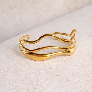 May include: A gold-colored cuff bracelet with a flowing, organic design. The bracelet features three interconnected bands, with a smooth, polished finish. The design has a wave-like appearance, adding a touch of elegance to the jewelry.