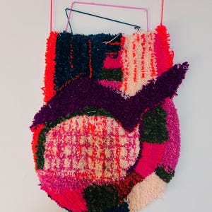 May include: Abstract textile artwork in a circular shape, featuring a mix of textures and colours. The piece includes shades of pink, purple, red, and green. It is suspended by a red string and a geometric frame, creating a modern aesthetic.